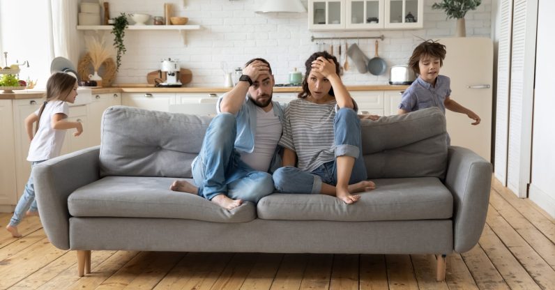 Tired mother and father sitting on couch feels annoyed exhausted while noisy little daughter and son shouting run around sofa where parents resting. Too active hyperactive kids, need repose concept